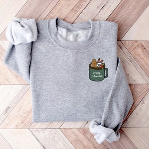 May include: A gray sweatshirt with a green mug design on the front. The mug is filled with a gingerbread man, marshmallows, and cinnamon sticks. The text on the mug reads "Merry Christmas".
