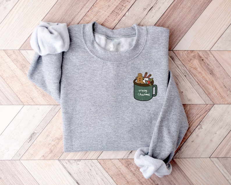 May include: A gray sweatshirt with a green mug design on the front. The mug is filled with a gingerbread man, marshmallows, and cinnamon sticks. The text on the mug reads "Merry Christmas".