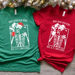 May include: Two Christmas themed t-shirts with a skeleton wearing a Santa hat and holding a mug of hot cocoa. The text on the shirts reads "When you're dead inside but it's Christmas". One shirt is green and the other is red.