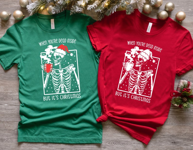 May include: Two Christmas themed t-shirts with a skeleton wearing a Santa hat and holding a mug of hot cocoa. The text on the shirts reads "When you're dead inside but it's Christmas". One shirt is green and the other is red.