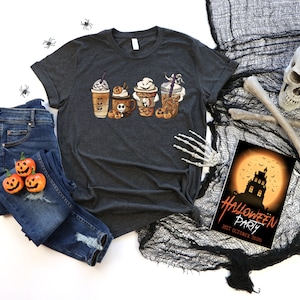 Pu&ograve; includere: Una maglietta grigio scuro con una grafica di quattro tazze da caff&egrave; a tema Halloween. Le tazze sono decorate con fantasmi, zucche e pipistrelli. La maglietta &egrave; appoggiata su una superficie bianca con un paio di jeans blu, un teschio e un invito a una festa di Halloween.