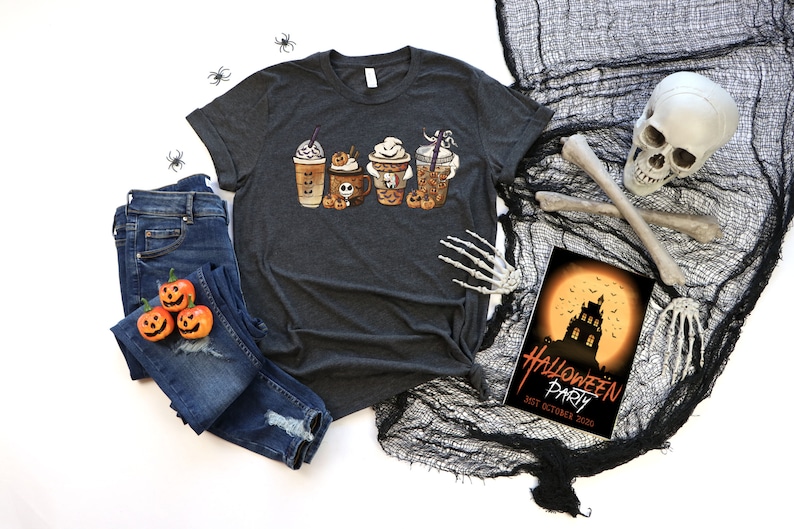 Pu&ograve; includere: Una maglietta grigio scuro con una grafica di quattro tazze da caff&egrave; a tema Halloween. Le tazze sono decorate con fantasmi, zucche e pipistrelli. La maglietta &egrave; appoggiata su una superficie bianca con un paio di jeans blu, un teschio e un invito a una festa di Halloween.