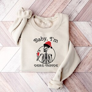 May include: A beige sweatshirt with a black and white skeleton wearing a red Santa hat and holding a red cup. The text "Baby, I'm Dead Inside" is printed on the sweatshirt.