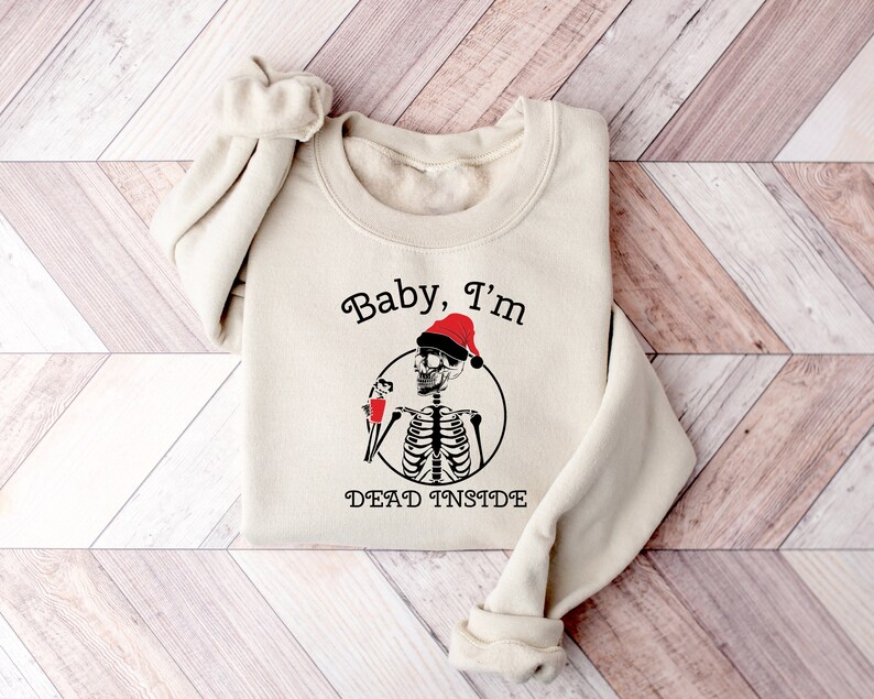 May include: A beige sweatshirt with a black and white skeleton wearing a red Santa hat and holding a red cup. The text "Baby, I'm Dead Inside" is printed on the sweatshirt.