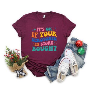 Puede incluir: Camiseta color burdeos con el texto "It's ok if your serotonin is store bought" en colores arco&iacute;ris.