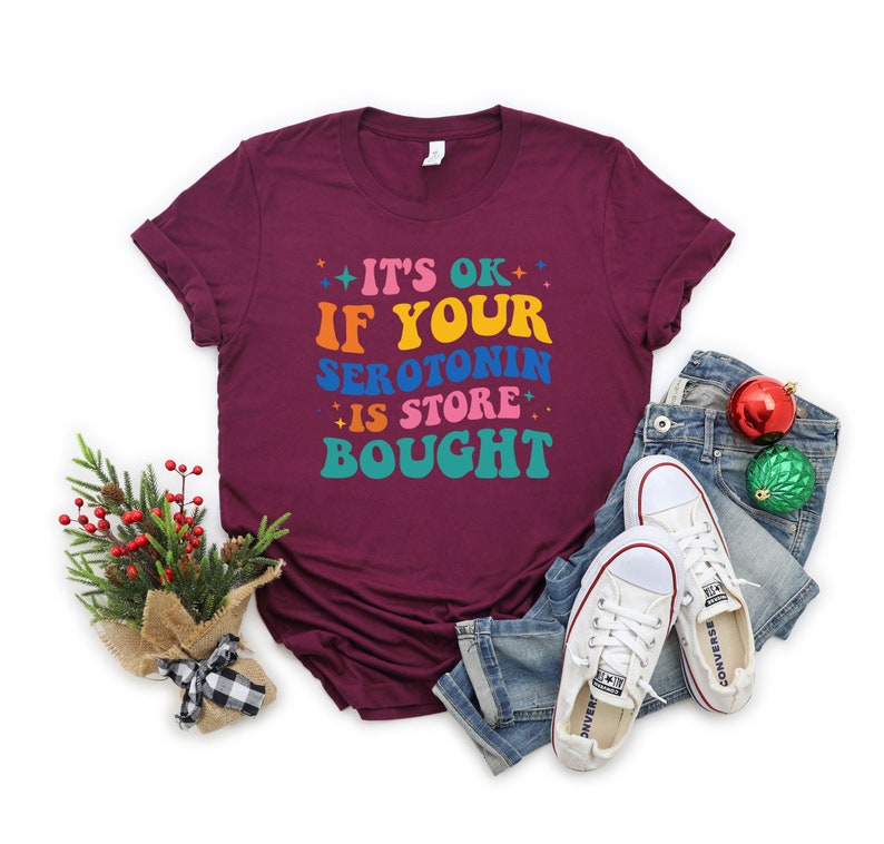 Puede incluir: Camiseta color burdeos con el texto "It's ok if your serotonin is store bought" en colores arco&iacute;ris.