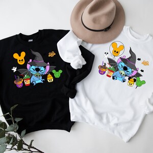 May include: Two sweatshirts featuring a blue Stitch character dressed as a witch holding a green alien. The Stitch character is surrounded by Halloween themed treats and decorations. One sweatshirt is black and the other is white.