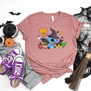 May include: A mauve t-shirt with a cartoon Stitch wearing a witch hat and holding a small ghost. The design also includes a Mickey Mouse balloon, a cauldron, and a Mickey Mouse shaped lollipop.