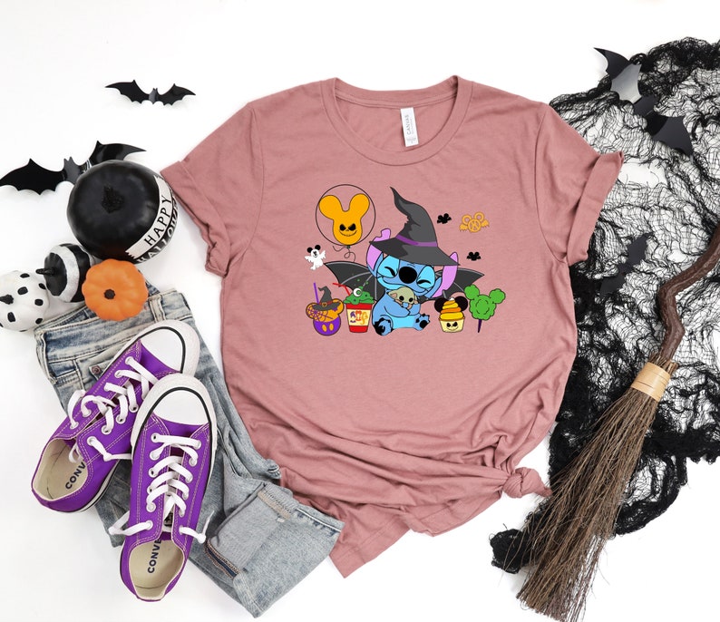 May include: A mauve t-shirt with a cartoon Stitch wearing a witch hat and holding a small ghost. The design also includes a Mickey Mouse balloon, a cauldron, and a Mickey Mouse shaped lollipop.