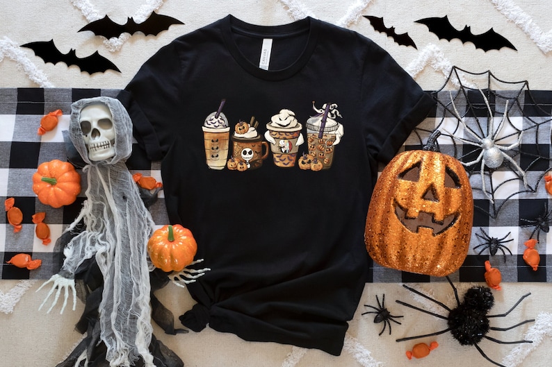 Pu&ograve; includere: Maglietta nera con una grafica di quattro tazze di caff&egrave; a tema Halloween. Le tazze sono decorate con fantasmi, zucche e altri disegni spettrali.