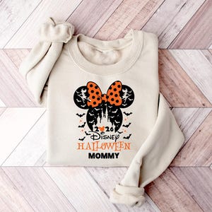 May include: A beige long-sleeve sweatshirt featuring a black and orange graphic of a Mickey Mouse silhouette with a polka dot bow, bats, and a castle. The text reads 2026 Disney HALLOWEEN MOMMY with a small orange heart between the numbers.