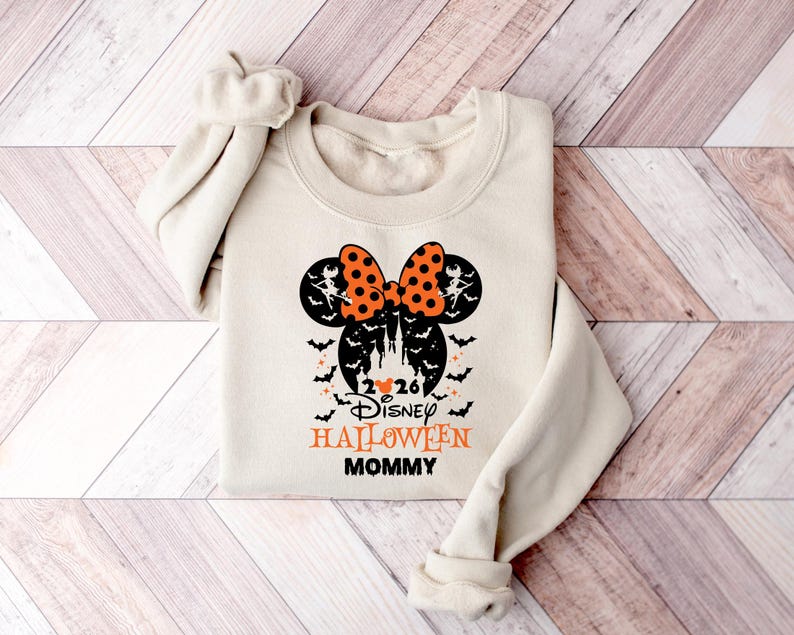 May include: A beige long-sleeve sweatshirt featuring a black and orange graphic of a Mickey Mouse silhouette with a polka dot bow, bats, and a castle. The text reads 2026 Disney HALLOWEEN MOMMY with a small orange heart between the numbers.