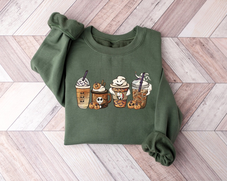 Pu&ograve; includere: Una felpa verde con una grafica di quattro bicchieri di caff&egrave; a tema Halloween con spezie di zucca e panna montata. I bicchieri sono decorati con Jack Skellington e altri personaggi di Halloween.