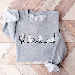 May include: A grey sweatshirt featuring a design of four cats dressed as ghosts. The cats are depicted in different poses, including standing, sitting, and lying down. The design is in black and white.