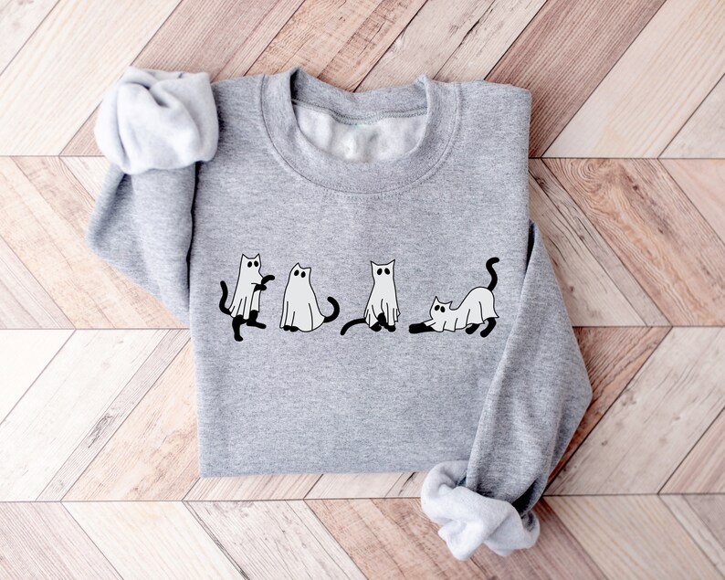 May include: A grey sweatshirt featuring a design of four cats dressed as ghosts. The cats are depicted in different poses, including standing, sitting, and lying down. The design is in black and white.