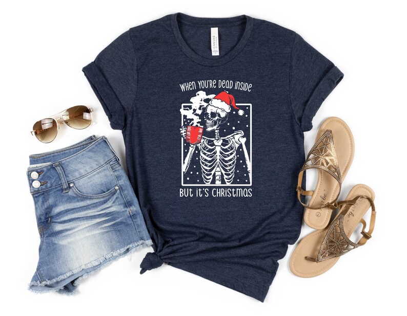 May include: A navy blue t-shirt with a white graphic of a skeleton wearing a Santa hat and holding a mug. The text on the shirt reads "When you're dead inside but it's Christmas".