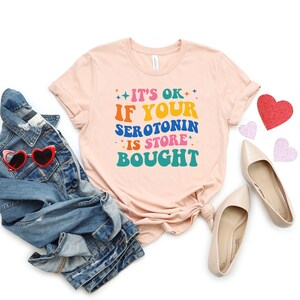 Puede incluir: Una camiseta rosa claro con el texto "It's ok if your serotonin is store bought" en letras de colores. La camiseta est&aacute; doblada y colocada sobre un fondo blanco con una chaqueta vaquera, gafas de sol y corazones rosas.