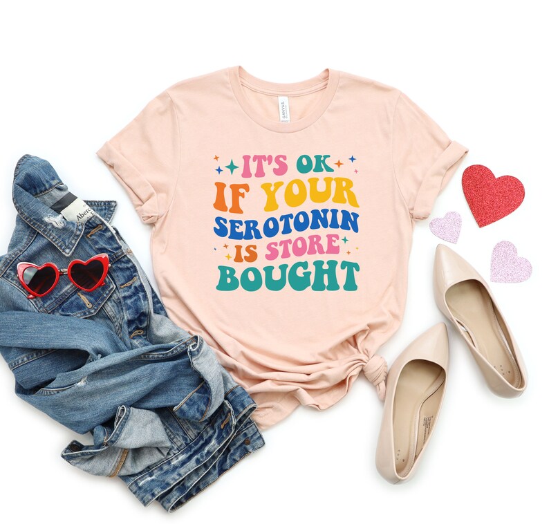 Puede incluir: Una camiseta rosa claro con el texto "It's ok if your serotonin is store bought" en letras de colores. La camiseta est&aacute; doblada y colocada sobre un fondo blanco con una chaqueta vaquera, gafas de sol y corazones rosas.