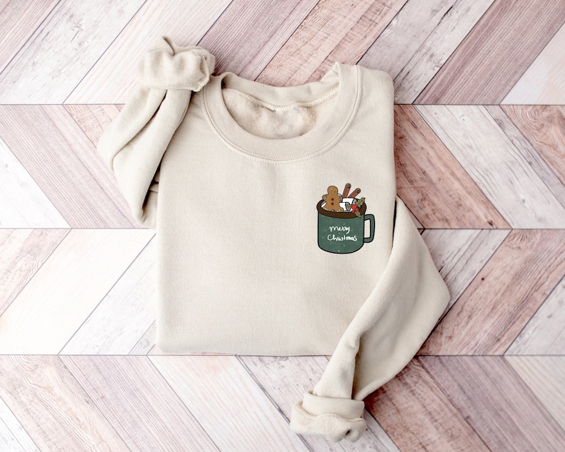 May include: A beige sweatshirt with a green mug design featuring a gingerbread man, marshmallows, and holly. The mug says "Merry Christmas".