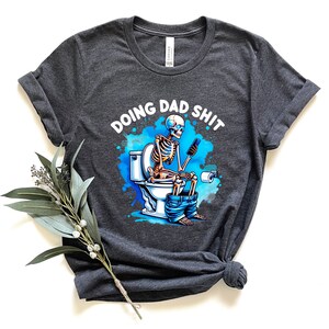 May include: Dark gray t-shirt with a humorous graphic. The design features a skeleton sitting on a toilet, using a phone, with the text "DOING DAD SHIT" above. The graphic has blue and white accents.