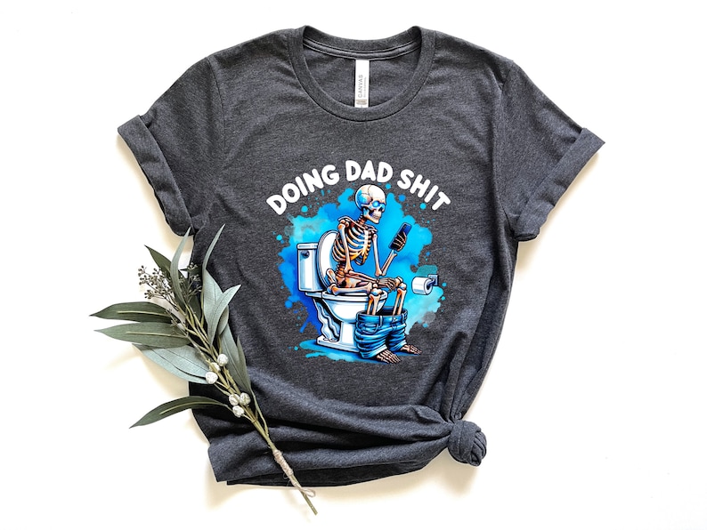 May include: Dark gray t-shirt with a humorous graphic. The design features a skeleton sitting on a toilet, using a phone, with the text "DOING DAD SHIT" above. The graphic has blue and white accents.