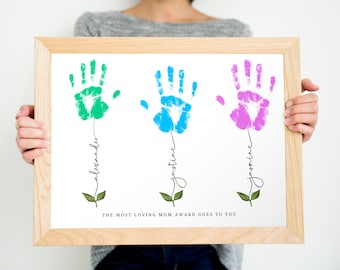 Puede incluir: Una impresión enmarcada con tres huellas de manos en verde, azul y rosa, cada una con un nombre escrito debajo: Alexander, Justine y Jasmine. El texto "The most loving mom award goes to you!" está en la parte inferior de la impresión.