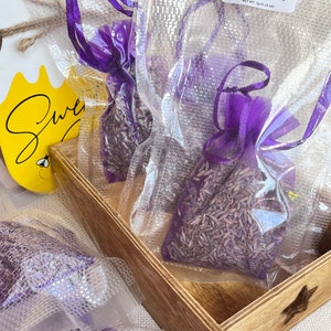 Può includere: Tre piccole borse di rete viola riempite di fiori di lavanda secchi. Le borse sono confezionate in sacchetti di plastica trasparente con etichette che recitano "Lavender Scent Bag" e "Net Wt. 7g (0.25 oz)".