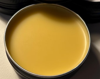May include: A close-up of a jar of Men's Beeswax Skin Balm in Bourbon Vanilla. The balm is a light yellow color and is in a black metal container. The label indicates the net weight is 2 oz.