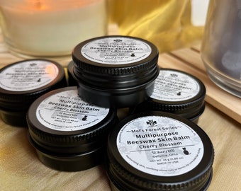 May include: Close-up of five small black metal tins with white labels. Each tin contains a "Multipurpose Beeswax Skin Balm" with a different scent. The scents are Cherry Blossom, Lavender, Eucalyptus, Tea Tree, and Frankincense. The tins are arranged in a circle on a gold fabric surface.