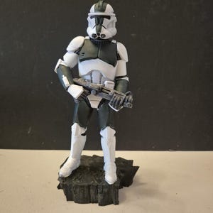 Pu&ograve; includere: Statuetta dettagliata di un clone trooper di Star Wars in posa eretta, con un fucile blaster. La figura &egrave; prevalentemente bianca e grigio scuro, con un elmetto e un'armatura. Si trova su una base scura e strutturata.