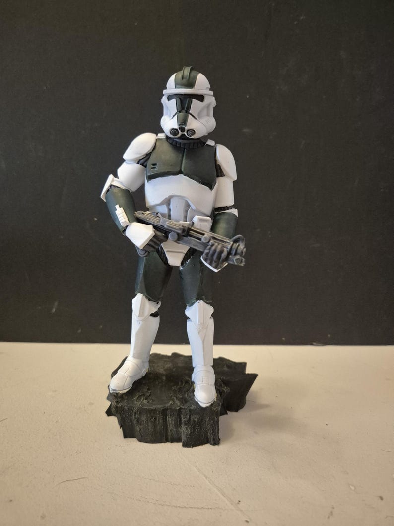 Pu&ograve; includere: Statuetta dettagliata di un clone trooper di Star Wars in posa eretta, con un fucile blaster. La figura &egrave; prevalentemente bianca e grigio scuro, con un elmetto e un'armatura. Si trova su una base scura e strutturata.