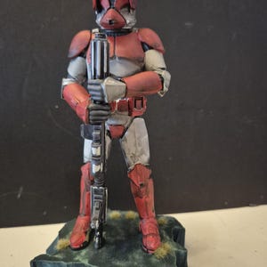 Pu&ograve; includere: Statuetta dettagliata di un soldato con armatura rossa e grigia, che tiene un fucile. La figura &egrave; su una piccola base strutturata con accenti verdi e marroni. L'elmo e l'armatura presentano dettagli intricati.