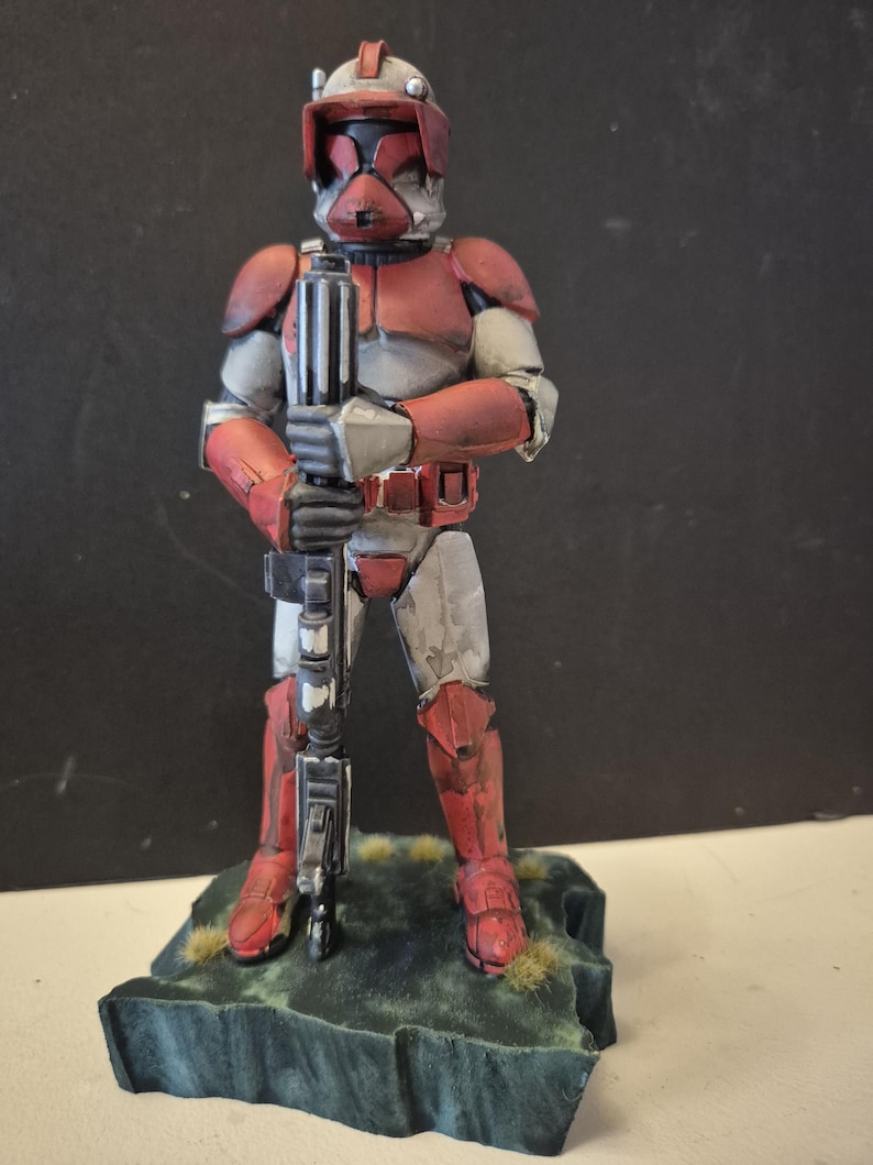 Pu&ograve; includere: Statuetta dettagliata di un soldato con armatura rossa e grigia, che tiene un fucile. La figura &egrave; su una piccola base strutturata con accenti verdi e marroni. L'elmo e l'armatura presentano dettagli intricati.