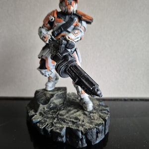 Puede incluir: Figura detallada de un soldado con armadura blanca y naranja, sosteniendo un arma negra grande. La figura se encuentra sobre una base rocosa y texturizada. El casco y la armadura tienen detalles intrincados.