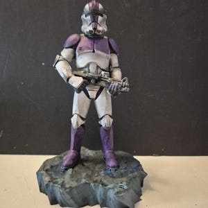 Pu&ograve; includere: Statuetta dettagliata di un soldato con armatura bianca e viola, che tiene un fucile blaster. La figura &egrave; su una base rocciosa grigia. L'elmo e l'armatura sono riccamente dettagliati.