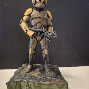 Pu&ograve; includere: Statuetta dettagliata di un soldato in tuta futuristica, principalmente nei toni del marrone e del giallo. La figura tiene un fucile e si trova su una base strutturata verde-marrone. L'armatura presenta dettagli intricati.