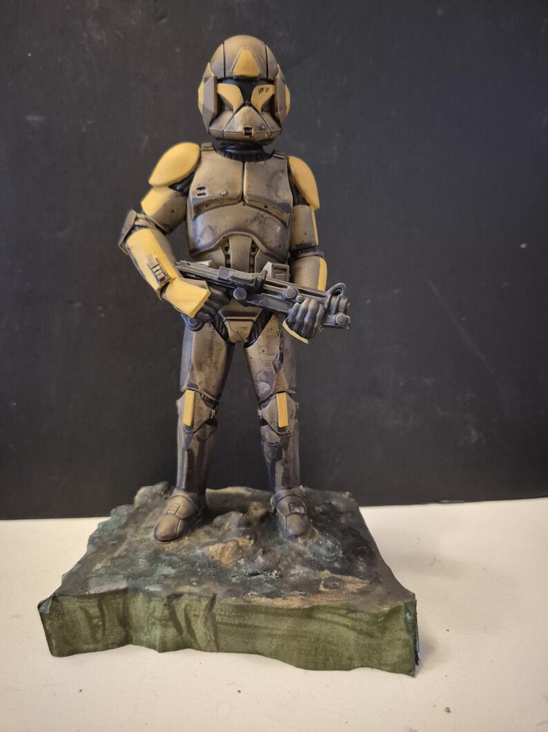 Pu&ograve; includere: Statuetta dettagliata di un soldato in tuta futuristica, principalmente nei toni del marrone e del giallo. La figura tiene un fucile e si trova su una base strutturata verde-marrone. L'armatura presenta dettagli intricati.