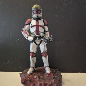 Pu&ograve; includere: Statuetta dettagliata di un clone trooper di Star Wars, con un fucile blaster. La figura &egrave; prevalentemente bianca con accenti bordeaux e grigi sul casco e sull'armatura. Poggia su una base strutturata marrone-rossastra.