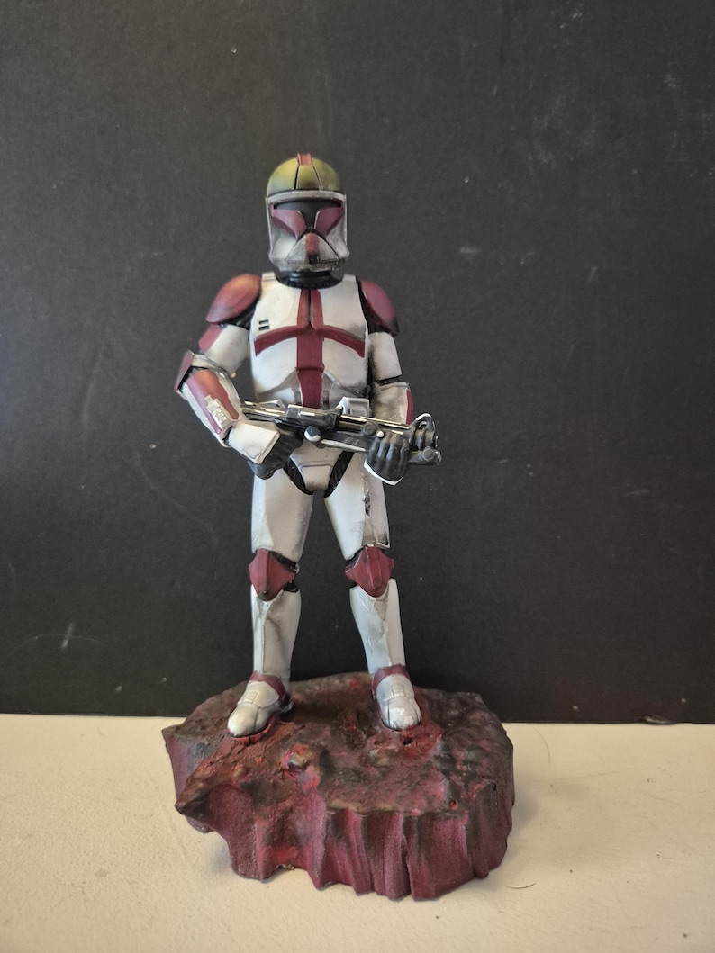 Pu&ograve; includere: Statuetta dettagliata di un clone trooper di Star Wars, con un fucile blaster. La figura &egrave; prevalentemente bianca con accenti bordeaux e grigi sul casco e sull'armatura. Poggia su una base strutturata marrone-rossastra.