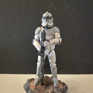 Pu&ograve; includere: Statuetta dettagliata di un soldato corazzato bianco e grigio, con un fucile. La figura &egrave; su una base rocciosa con toni marroni e grigi. L'elmo e l'armatura del soldato sono progettati in modo intricato, mostrando un'estetica futuristica.