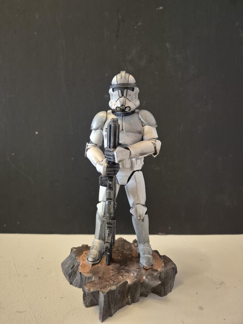 Pu&ograve; includere: Statuetta dettagliata di un soldato corazzato bianco e grigio, con un fucile. La figura &egrave; su una base rocciosa con toni marroni e grigi. L'elmo e l'armatura del soldato sono progettati in modo intricato, mostrando un'estetica futuristica.