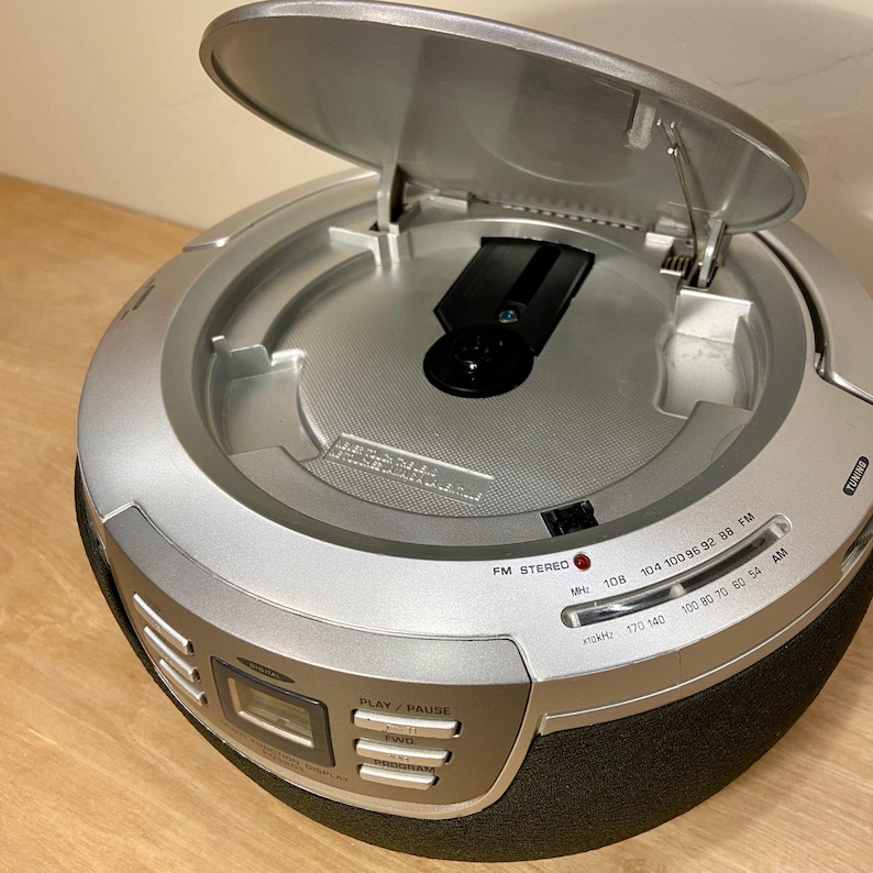 May include: Silver and black portable CD player with the CD tray open. The player has a digital display, FM radio tuner, and control buttons. The top of the player is silver, and the bottom is black. The radio tuner displays frequencies in MHz and kHz.
