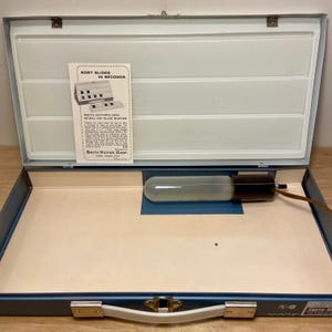 May include: An open, light blue metal case with a white interior, containing a long, cylindrical light bulb and a printed card. The card reads "SORT SLIDES IN SECONDS" and details the Smith-Victor Model 551 Slide Sorter.