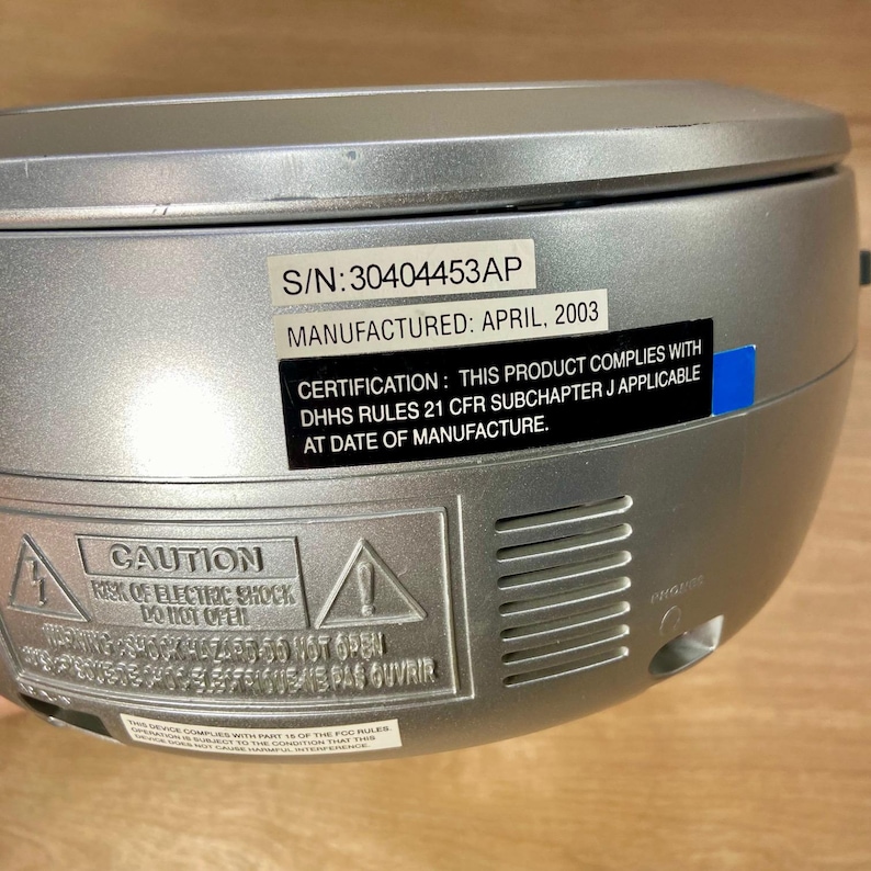 May include: Silver-colored portable CD player with a curved top. The device has several labels with text, including "S/N: 30404453AP" and "MANUFACTURED: APRIL, 2003." A warning label reads "CAUTION RISK OF ELECTRIC SHOCK DO NOT OPEN."