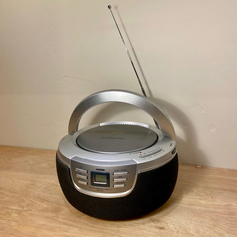 May include: A silver and black portable CD player with a handle and an antenna. The CD player has a digital display and control buttons. The speaker portion is black and the top is silver.
