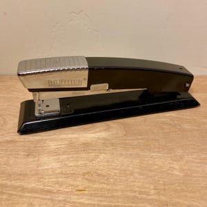 May include: A black and silver Bostitch stapler. The stapler has a black body with a silver top and the brand name "BOSTITCH" printed on the side. The stapler is sitting on a wooden surface.