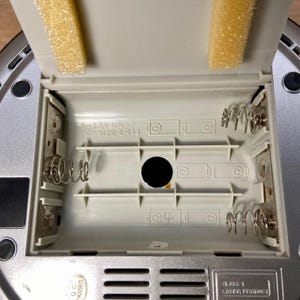 May include: An open battery compartment of a silver and gray electronic device. The compartment is empty, revealing spring contacts and slots for batteries. The lid has yellow foam padding. Text on the device reads "5x1.5V UN-2 C SIZE LR14".