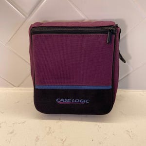 May include: A rectangular Case Logic storage pouch featuring a deep purple upper section and a black lower section, separated by a thin blue horizontal stripe. The black section displays the Case Logic brand name in blue text with a zipper closure.