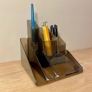 May include: A clear brown desk organizer with multiple compartments. It holds a blue pen, yellow pencils, and black markers. The organizer has a tiered design and a small tray at the base.