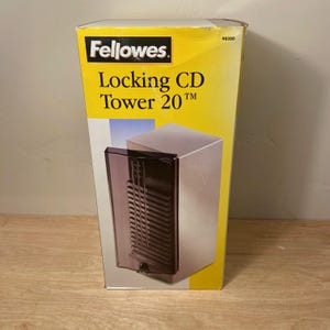May include: A Fellowes Locking CD Tower 20 product box with a yellow and white background. The box features a graphic of a silver storage tower with a dark, translucent, hinged door and a small lock mechanism at the bottom center of the unit.